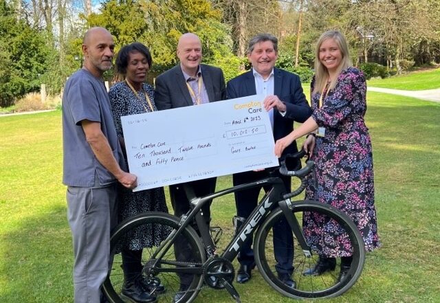 Image description: 5 people stand behind a bike, in the middle of a garden, smiling and holding a giant cheque made out to Compton Care for £10,012.50