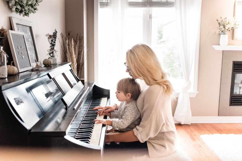 A person with long blonde hair is sat playing a piano with a small child on their lap, watching their hands